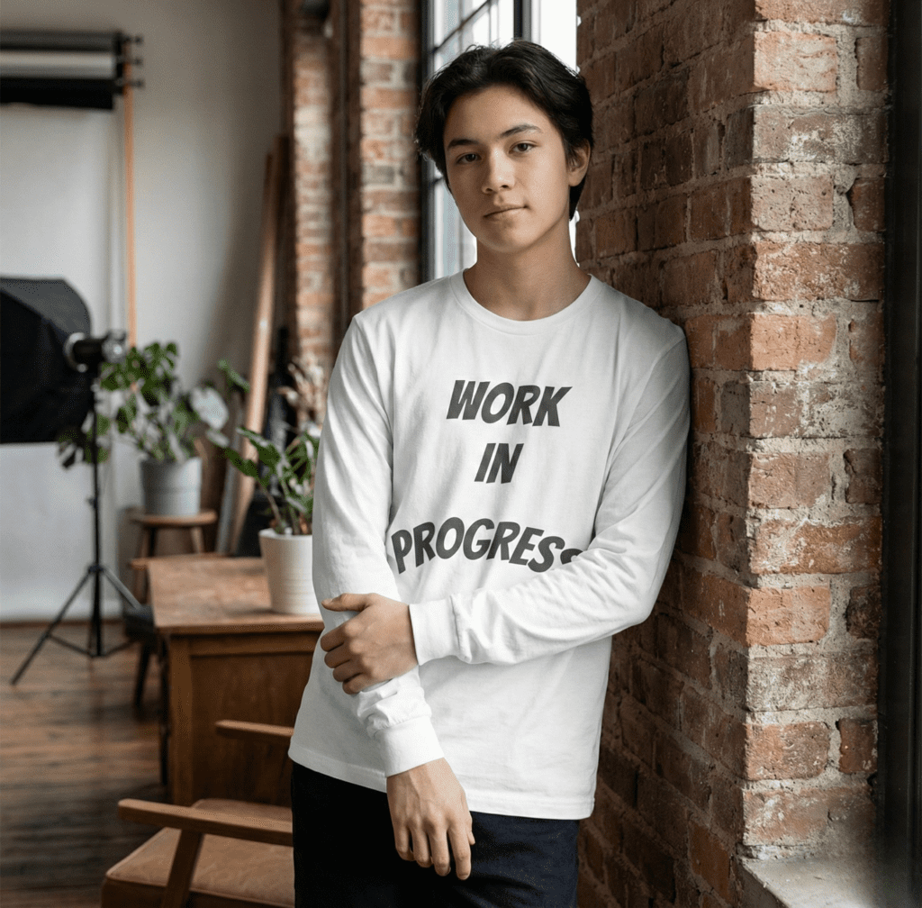 A young man leaning against a brick wall wearing a white Work in Progress Graphic Long Sleeve Tee with bold grey arched text.
