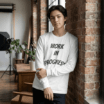 A young man leaning against a brick wall wearing a white Work in Progress Graphic Long Sleeve Tee with bold grey arched text.