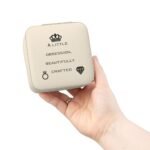 A person's hand holding a high-quality, cream-colored Premium Jewelry Storage Box featuring a black crown icon and the quote “A Little Obsession, Beautifully Crafted.”